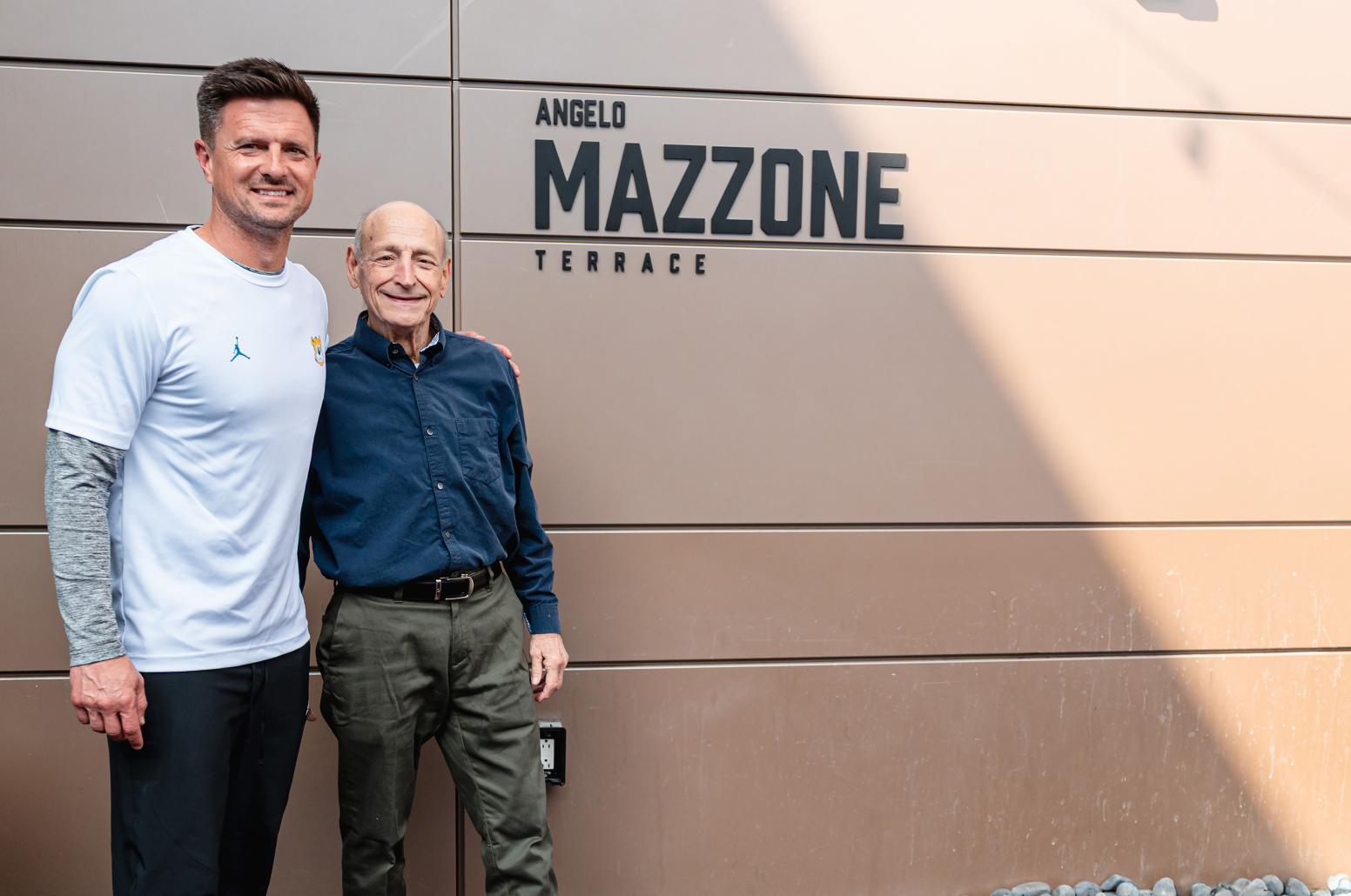 Bob Chesney (left), in white T-shirt and black pants standing next to Angelo Mazzone, in dark blue button-down and gray slacks, next to Angelo Mazzone Terrace sign