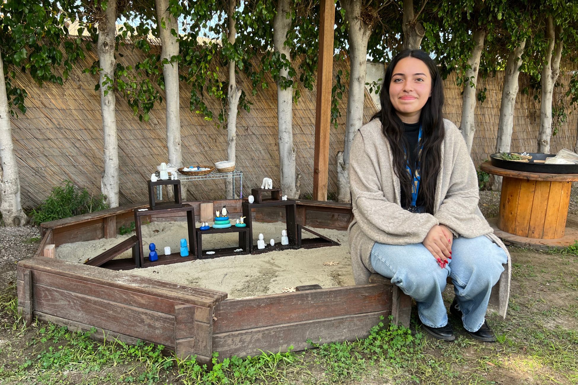 A young woman sits on the edge of a sandbox. She wears long black hair, a slight smile, jeans and tan cardigan.