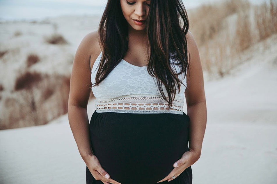 A dark haired pregnant woman is looking down and touching her belly at the beach.